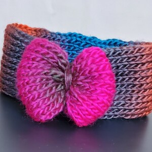 May include: A knitted headband with a large pink bow. The headband is made with a multi-colored yarn in shades of orange, blue, and black.