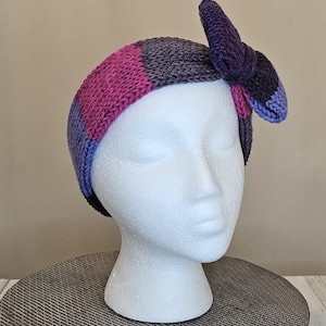 Puede incluir: Una diadema de punto a rayas de color morado, rosa y azul con un gran lazo en un lado.