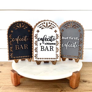 May include: Three decorative wooden signs with arched tops, each with a different color and text. The signs are black, white, and gray, and say "cafecito BAR", "cafecito y chisme BAR", and "but first, cafecito". They sit on a white marble stand.