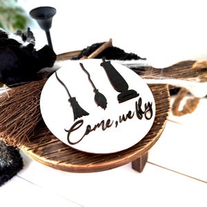 May include: A round, white wooden sign with the phrase "Come, we fly" in script, featuring black silhouettes of a broom, a witch's broom, and a vacuum cleaner. The sign is on a small, wooden stand with a rustic finish.