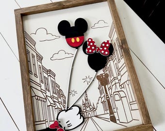 May include: Wooden framed art piece featuring a black and white street scene with Mickey and Minnie Mouse balloon designs. The Mickey balloon has red accents, and the Minnie balloon has a red polka dot bow. The frame is a natural wood color.