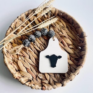 May include: A decorative farmhouse-style wooden tag with a black cow head silhouette, strung with speckled beads. The tag is white and sits in a woven basket with wheat stalks. The overall color palette is neutral with natural textures.