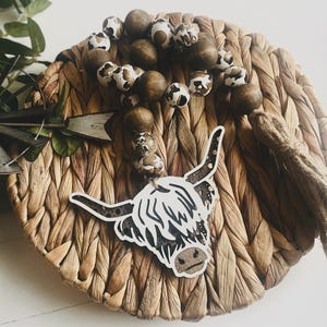 May include: A decorative beaded garland with a Highland cow charm. The garland features alternating brown and white patterned beads and solid brown beads. The cow charm is black and white with a brown nose and ears. The garland is displayed on a woven basket.
