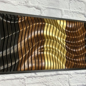 May include: A wall-mounted wood sculpture with a wavy pattern in black, brown, and gold tones. The sculpture is framed in a black wood frame.