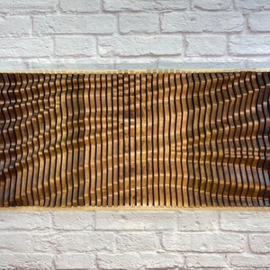 May include: A wall art piece made of wood with a wavy pattern. The wood is a dark brown color and the piece is mounted on a white brick wall.