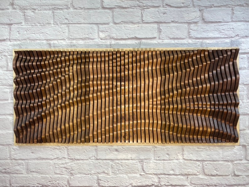 May include: A wall art piece made of wood with a wavy pattern. The wood is a dark brown color and the piece is mounted on a white brick wall.