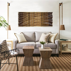 May include: A living room with a gray striped sofa, two wooden end tables, a black metal chair, and a wooden wall sculpture with a wavy pattern. The room has a light brown rug and white walls.