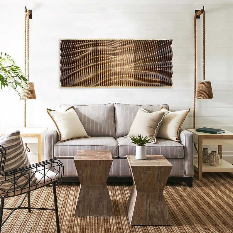 May include: A living room with a gray striped sofa, two wooden end tables, a black metal chair, and a wooden wall sculpture with a wavy pattern. The room has a light brown rug and white walls.