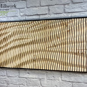 May include: A wooden wall art piece with a wavy pattern, framed in black. The piece is made of thin strips of wood that are arranged in a wave-like pattern. The wood is a light brown color. The piece is titled "Art Millwork made in USA".