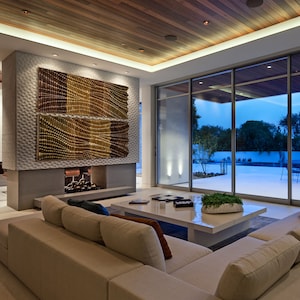 May include: A modern living room with a white sectional sofa, a white coffee table, and a large window overlooking a patio with a pool. The room features a fireplace with a decorative metal screen and a large abstract wall art piece.