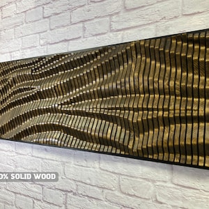 Puede incluir: Una gran escultura de pared abstracta hecha de madera maciza. La escultura está compuesta por muchas piezas individuales de madera que están dispuestas en un patrón de onda. La madera es de color marrón oscuro y tiene un acabado natural. La escultura está enmarcada en un marco negro. El texto "100% SOLID WOOD" está impreso debajo de la escultura.