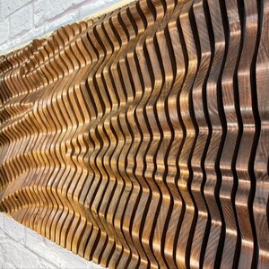 May include: A wall sculpture made of wood with a wavy pattern. The wood is a light brown color and has a natural grain. The sculpture is made up of many individual pieces of wood that are stacked on top of each other.