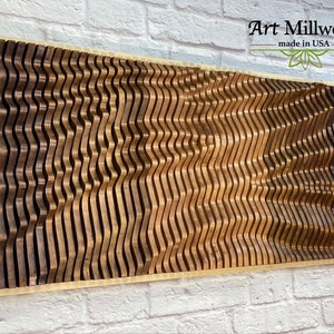 May include: A wooden wall art piece with a wavy pattern in shades of brown. The piece is framed in a light wood frame and has the text "Art Millwork - made in USA" in the top right corner.