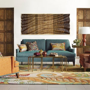 May include: A teal blue sofa with colorful patterned pillows sits in a living room with a large wooden wall art piece above it. The sofa is in front of a colorful rug with a patterned design. There are two gold metal coffee tables in front of the sofa.
