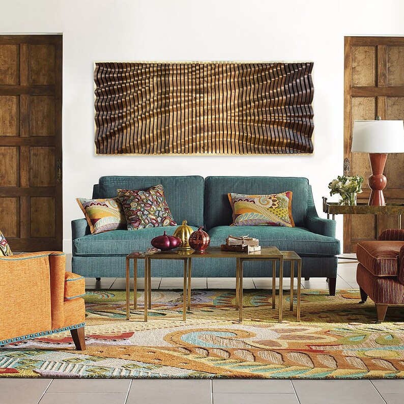 May include: A teal blue sofa with colorful patterned pillows sits in a living room with a large wooden wall art piece above it. The sofa is in front of a colorful rug with a patterned design. There are two gold metal coffee tables in front of the sofa.