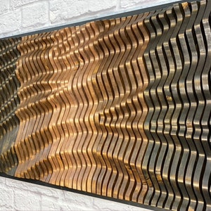 May include: A wall art piece made of wood with a wavy pattern. The wood is stained in three different shades: black, brown, and a lighter brown. The piece is mounted on a black frame.