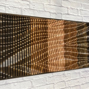 Op de afbeelding: Een aan de muur gemonteerd houten kunstwerk met een golvend patroon. Het stuk is gemaakt van meerdere dunne stroken hout in verschillende bruintinten, waardoor een 3D-effect ontstaat.