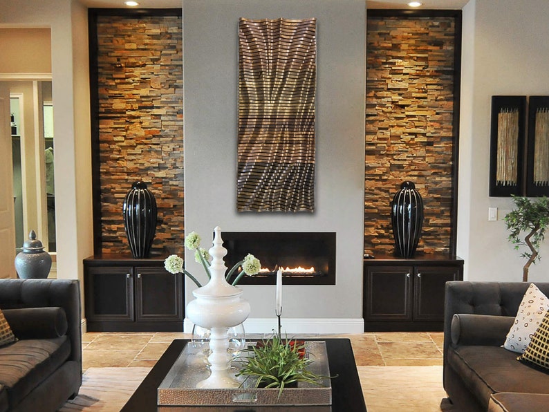 May include: A modern living room with a fireplace and a metal wall sculpture. The fireplace is surrounded by stone and the sculpture is a geometric design in shades of gold and brown.