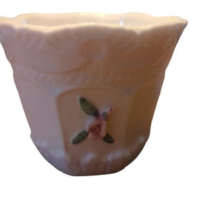 May include: A pale pink ceramic planter with a hexagonal shape and a scalloped rim. The planter features embossed floral detailing and a painted rose with green leaves. This decorative planter is suitable for small plants.