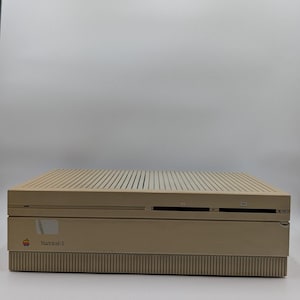 May include: A beige Macintosh II computer case featuring a horizontal rectangular design with a vented top panel, two front-facing disk drive slots, and a small colorful Apple logo next to the text Macintosh II printed on the front panel.