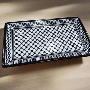 May include: Rectangular black and white ceramic serving tray. The tray features a geometric pattern of black diamonds on a white background. The border has a zig-zag design. The tray is on a light brown wooden surface.