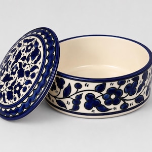 May include: A round ceramic container with a matching lid, featuring a cream-colored base decorated with hand-painted dark blue floral and vine patterns. The lid is tilted to the side, revealing the smooth, plain interior of the circular vessel.