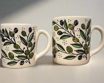 May include: Two off-white ceramic mugs of different sizes, each featuring a hand-painted pattern of green olive branches with black olives and small black dots. Both mugs have a cylindrical body and a curved handle on the right side.