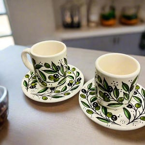 Puede incluir: Dos tazas de espresso de cerámica color hueso con platos a juego, decoradas con un patrón pintado a mano de ramas de olivo verdes y negras. Cada taza presenta un asa curva y una forma cilíndrica, mientras que los platos muestran un motivo.