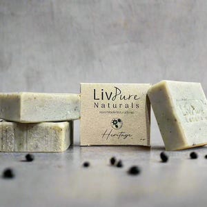 Puede incluir: Tres pastillas de jabón rectangulares de color claro con motas. Una pastilla está envuelta en una etiqueta de papel color canela con el texto impreso LivPure Naturals, Hand Made Natural Soap y Heritage. Dos pastillas están apiladas.