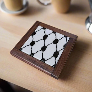 May include: Square wooden coaster with a white tile insert featuring a black dot and line pattern. The coaster has a dark brown wooden frame. The coaster is on a light brown wooden surface.