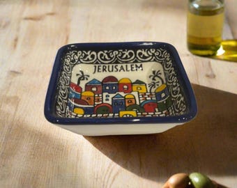May include: A square ceramic bowl with a dark blue rim and a hand-painted scene of colorful buildings and palm trees. The word JERUSALEM is printed in black at the top center. The interior border features a decorative black and white pattern.
