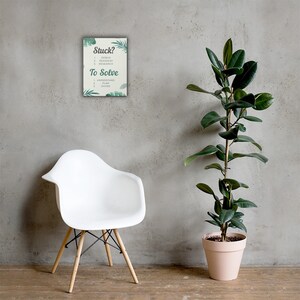 May include: A framed print with the text "Stuck?" and "To Solve" with a list of steps. The print is decorated with green leaf illustrations. A white chair with wooden legs is in front of a gray wall. A tall potted plant is to the right.