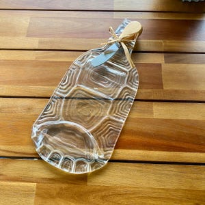 May include: A clear, glass serving platter in the shape of a bottle, with a textured surface. A small, gray spreader is tied to the top with a natural-colored ribbon. The platter is on a wooden surface.