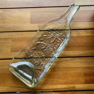Può includere: Un vassoio da portata in vetro trasparente a forma di bottiglia di vino con un design testurizzato di api e fiori. Il vassoio è su una superficie di legno.