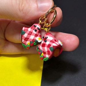 May include: A pair of gold hoop earrings with whimsical, heart-shaped charms. The charms feature a red and white checkered pattern, adorned with miniature strawberries, flowers, and green leaves. The earrings are a playful accessory.