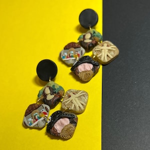 May include: A pair of dangle earrings with multiple charms. The charms feature various images, including a person, a group of people, and a tree. The earrings are made of polymer clay and have a black base.