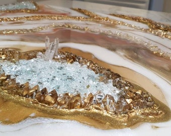 Puede incluir: Una pieza de arte de resina blanca y dorada con una gran geoda de cristal transparente en el centro. La geoda está rodeada de purpurina dorada y cristales más pequeños.