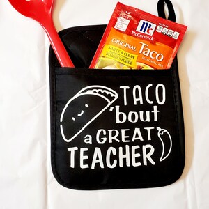 May include: Black oven mitt with a white taco graphic and the text "Taco 'bout a Great Teacher" and a red chili pepper. A red slotted spoon and a packet of McCormick Original Taco Seasoning Mix are in the pocket of the oven mitt.