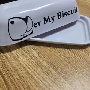 May include: A white butter dish with a lid, featuring the text "er My Biscuit" and a graphic of a rear end with a heart. The dish is open, revealing the interior. The design is in black.