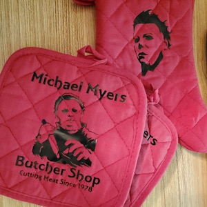 May include: A set of two red quilted oven mitts with black text and images. One mitt features a black and white image of Michael Myers with the text "Michael Myers Butcher Shop Cutting Meat Since 1978". The other mitt features a black and white image of Michael Myers's face.