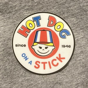 May include: A white circle sticker with a red, yellow, and blue logo. The logo says "Hot Dog On A Stick" and "since 1946". The logo features a cartoon person wearing a red, white, and blue hat.