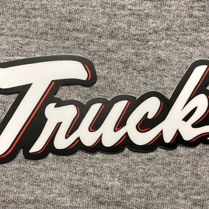 May include: White vinyl sticker with the word "Truck" in a black outline with a red inner outline. The letters are in a stylized script font.