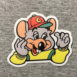 May include: A cartoon sticker of a gray mouse wearing a red and green baseball cap with the letter "C" on it. The mouse is smiling and has a yellow shirt with a green collar. The mouse is waving with its right hand.
