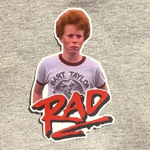 May include: A red and white sticker with a photo of a young boy with red hair wearing a white t-shirt with the text "Bart Taylor" and a graphic of a person riding a motorcycle. The text "RAD" is in large red letters with a black outline.