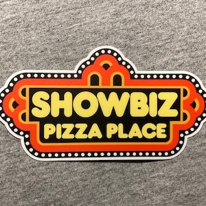 May include: A red and white sign with a black background and white polka dots around the border. The sign says "Showbiz Pizza Place" in yellow letters.