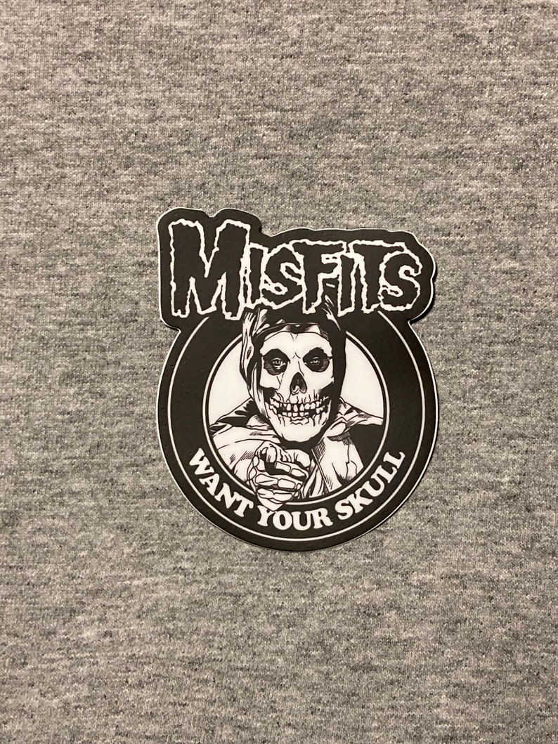 May include: Black and white sticker with the band name "Misfits" in bold letters. The sticker features a skull-faced character with a pointed finger pointing towards the viewer. The text "Want Your Skull" is written in a circle around the character.