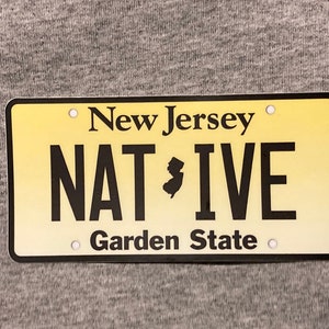 Pu&ograve; includere: Targa rettangolare con sfondo sfumato giallo. Il testo "New Jersey" &egrave; in alto, con "NATIVE" in grandi lettere nere e una silhouette del New Jersey. "Garden State" &egrave; in basso.