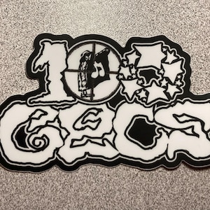 May include: A black and white sticker with the text "100 GECS" in a stylized font. The design includes a crosshair with a figure inside, and a cluster of stars. The sticker has a black border.