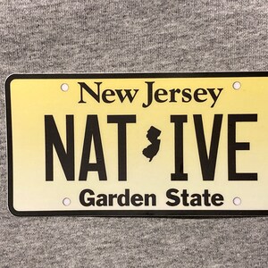 Pu&ograve; includere: Targa rettangolare del New Jersey con sfondo sfumato giallo. La targa presenta le parole "New Jersey" in alto, "NATIVE" in grassetto nero e "Garden State" in basso. Una silhouette nera dello stato &egrave; al centro.