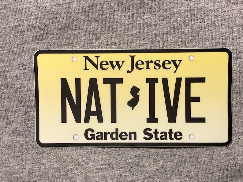 Pu&ograve; includere: Targa rettangolare del New Jersey con sfondo sfumato giallo. La targa presenta le parole "New Jersey" in alto, "NATIVE" in grassetto nero e "Garden State" in basso. Una silhouette nera dello stato &egrave; al centro.